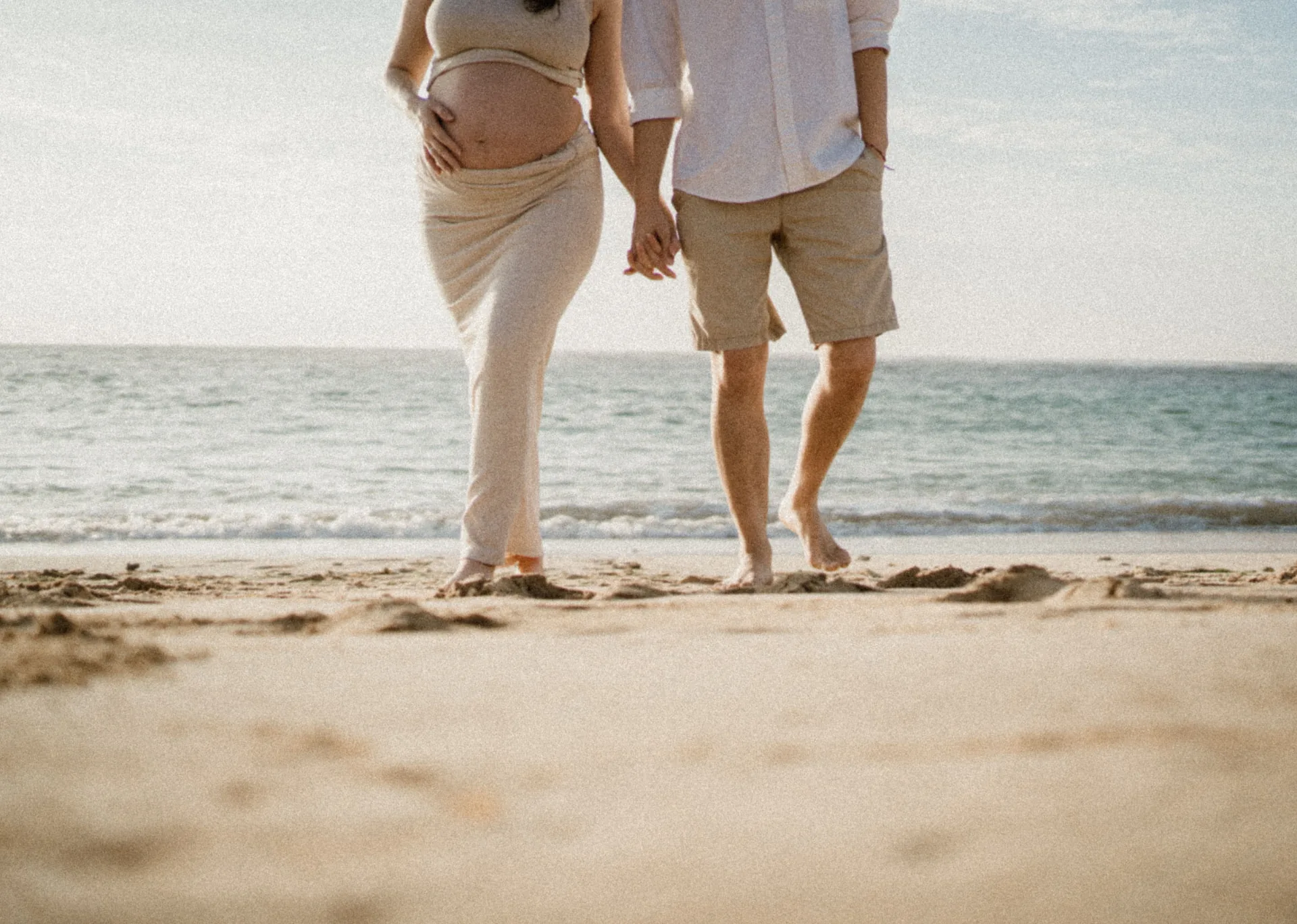 Expectant couple holding hands while walking along the shoreline at golden hour