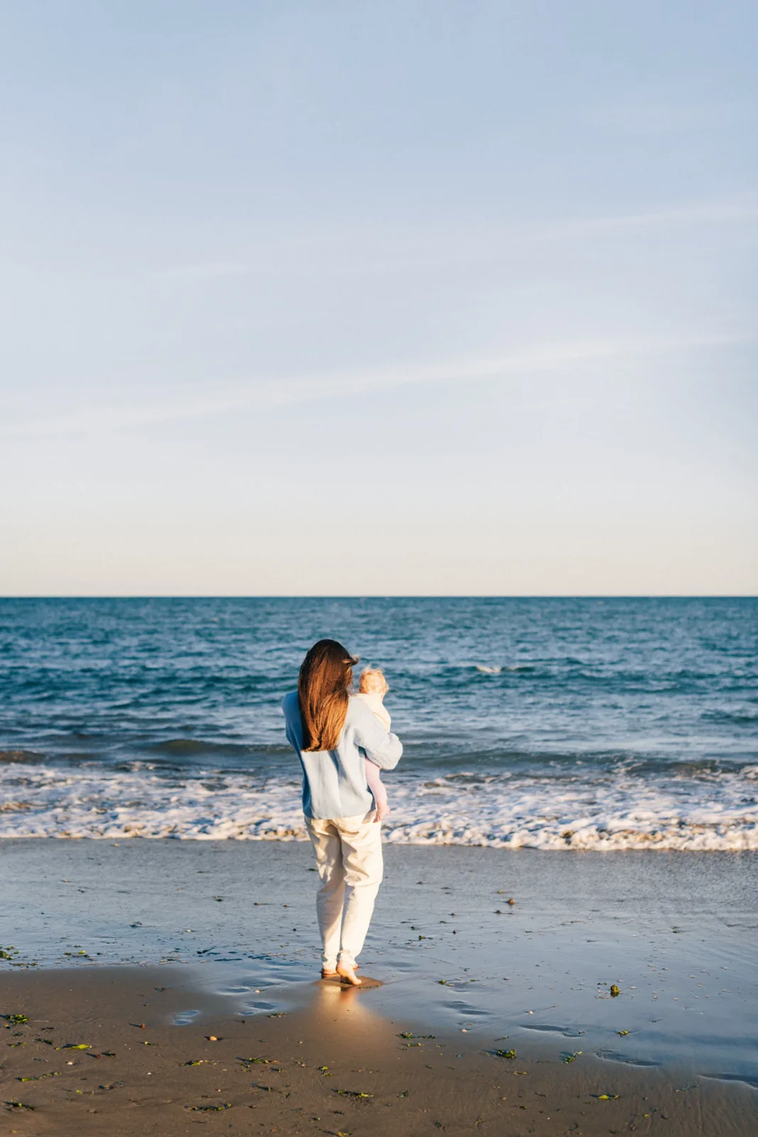 Mother holding her baby, facing the ocean at the water's edge
